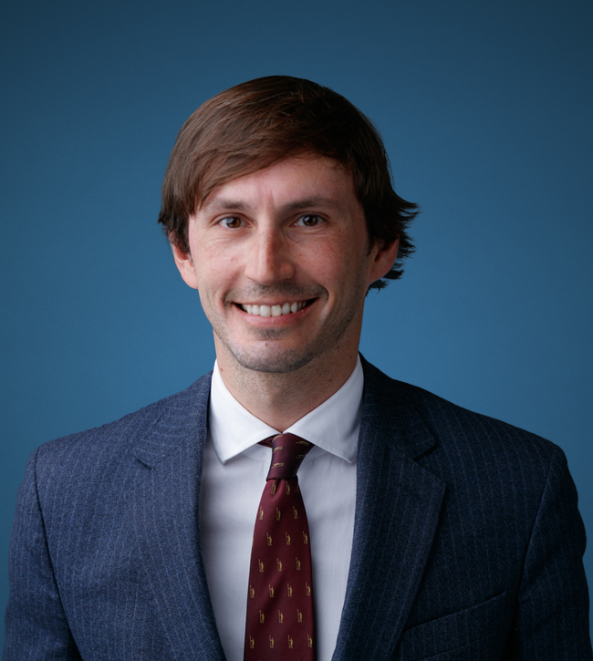 daniel-romagnolo_ Smiling man in a navy pinstripe suit and white shirt against a blue backdrop, professional headshot.