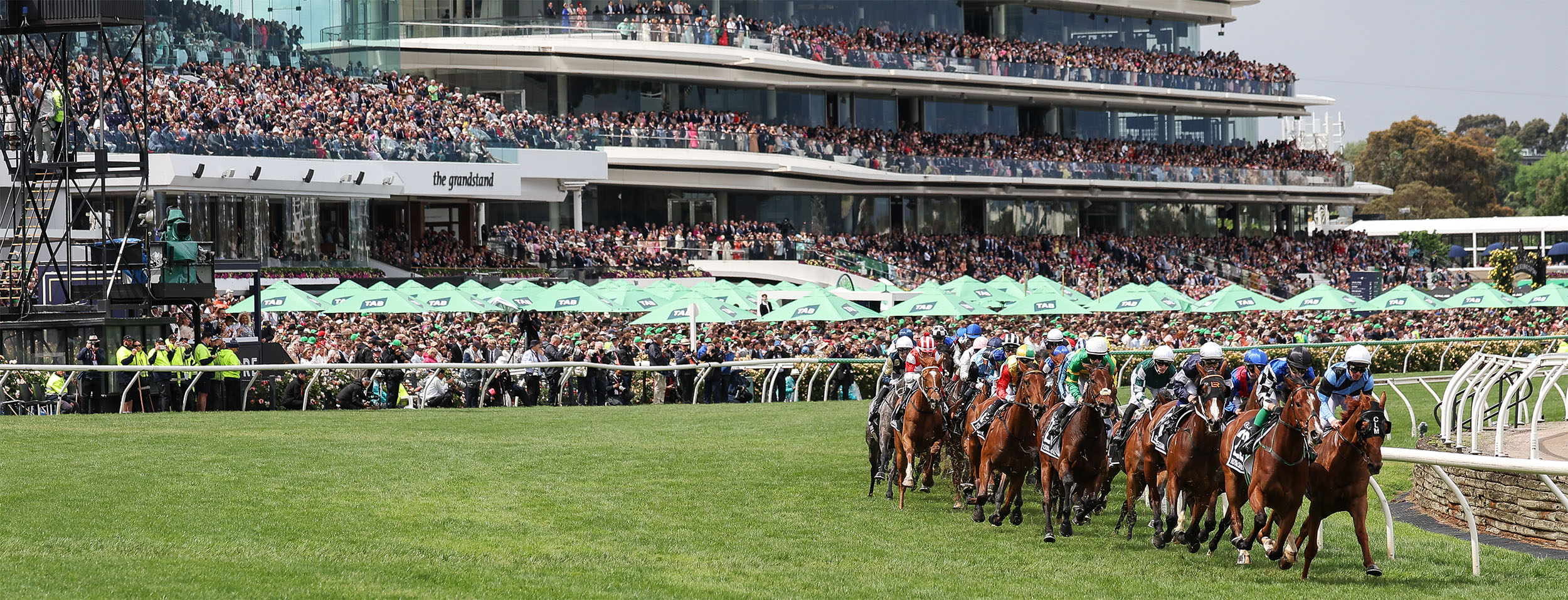 new-victoria-racing-club Horses with jockeys in colorful silks race along a grassy track, a packed grandstand visible above with spectators and green umbrellas in the infield.