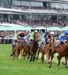 VRC-official-banner Jockeys on horses race along a grassy track with a packed grandstand in the background and spectators under umbrellas.